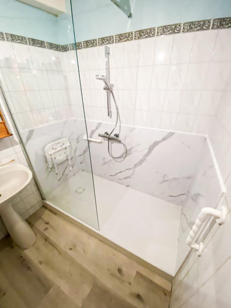 Douche à l'italienne moderne et accessible: carrelage blanc, parois marbrées, siège pliable, barre d'appui. Sol effet bois.