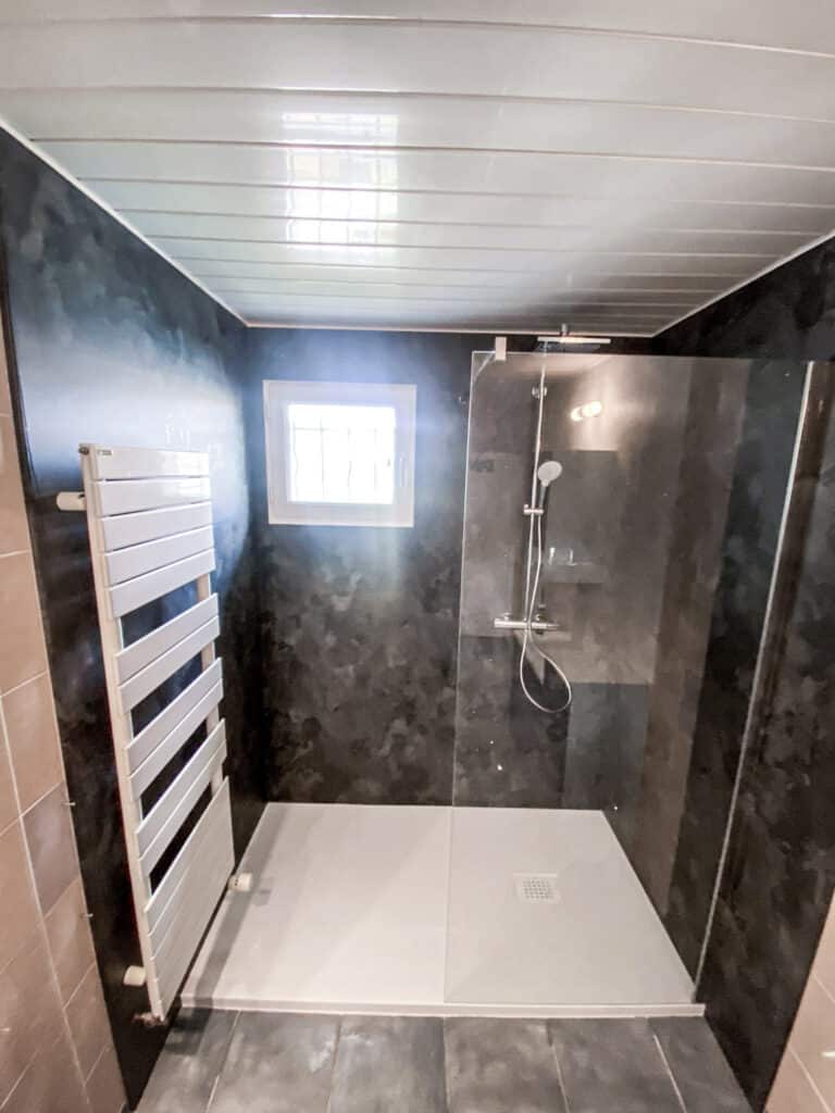 Salle de bain moderne avec douche à l'italienne, murs sombres, plafond à lattes clair. Radiateur sèche-serviettes blanc et fenêtre.