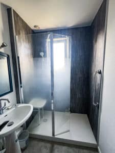 Salle de bain moderne avec douche à l'italienne, parois vitrées, panneaux muraux gris foncé. Lavabo blanc, siège de douche, fenêtre lumineuse.