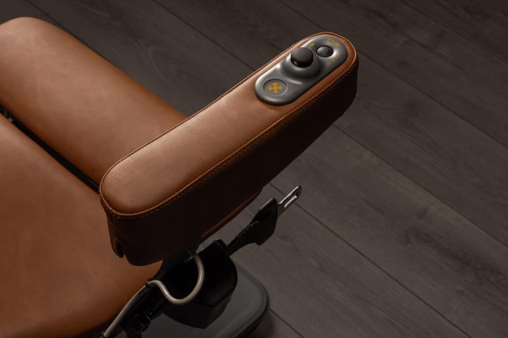 Accoudoir en cuir marron de fauteuil motorisé. Commandes : joystick, boutons "STOP", micro, marche/arrêt, alerte et croix. Ceinture de sécurité.