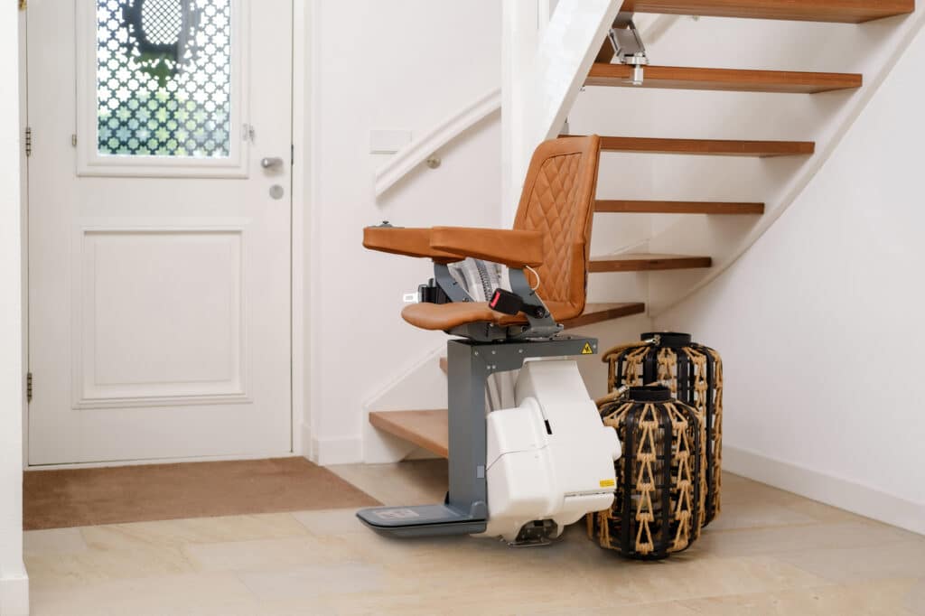 Monte-escalier moderne avec siège en cuir matelassé marron, installé près d'un escalier en bois et d'une porte blanche dans un intérieur lumineux.