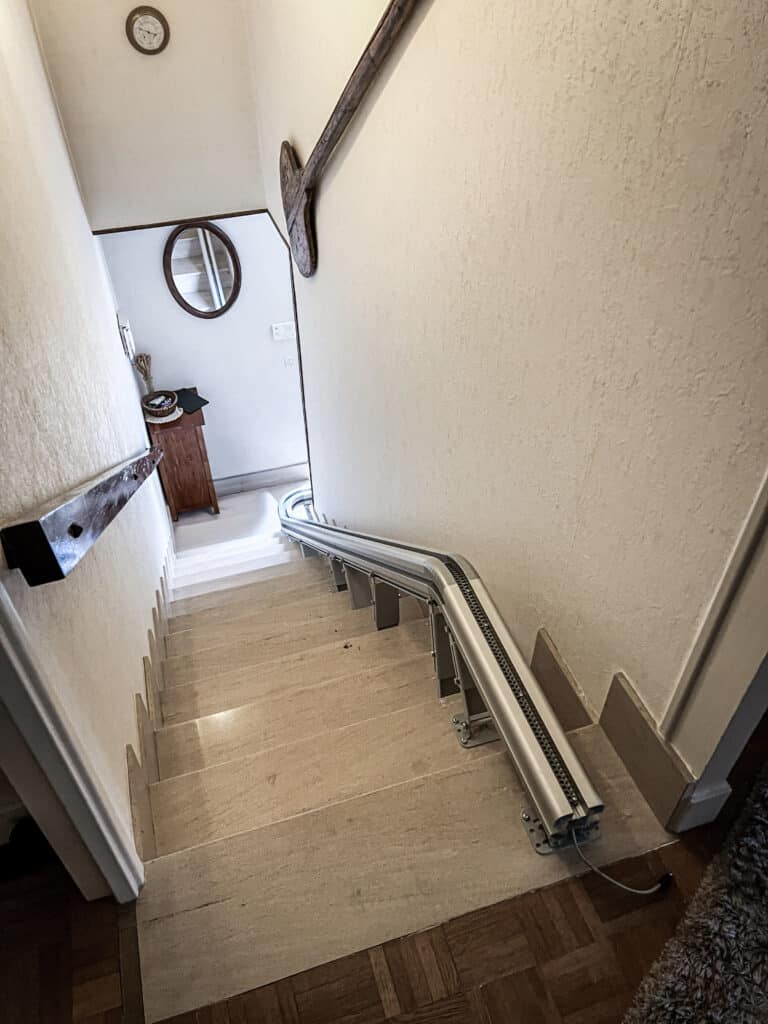 Escalier intérieur beige avec rail de monte-escalier métallique à droite. Miroir rond et horloge au palier supérieur, main courante en bois à gauche.