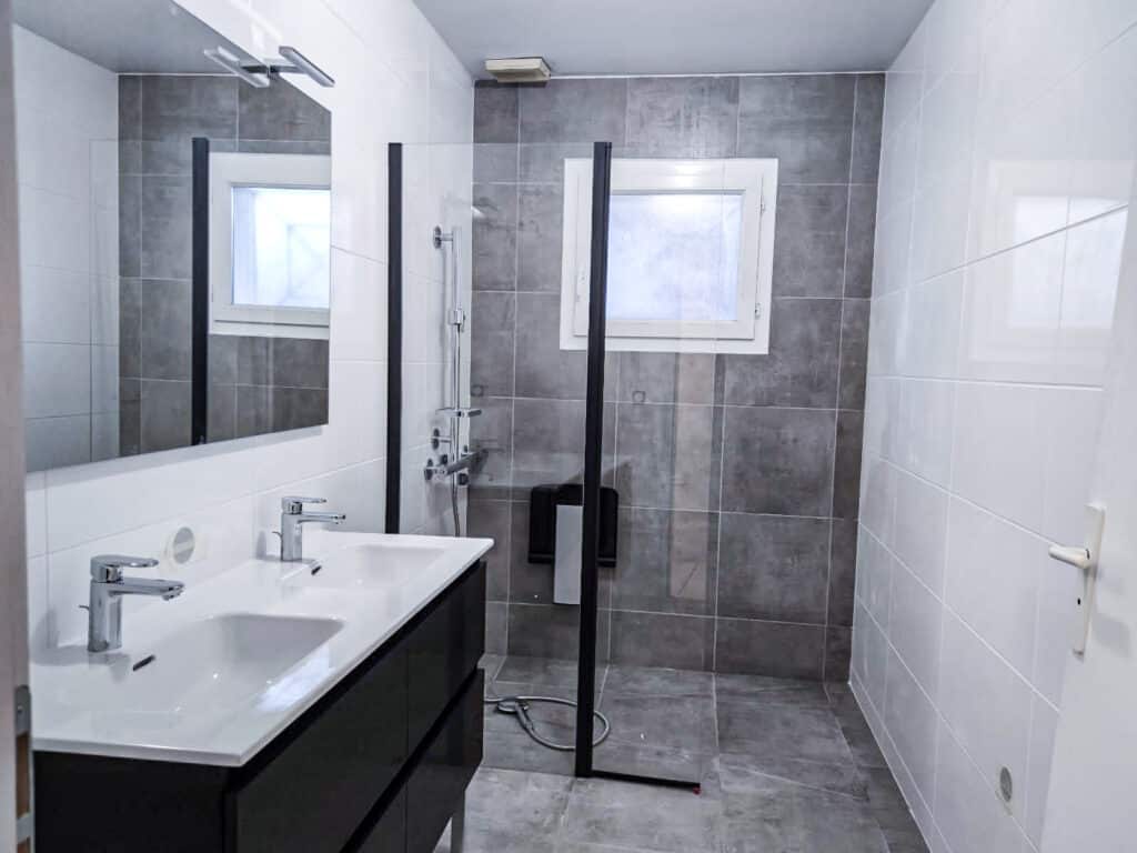 Salle de bain moderne: double vasque & douche à l'italienne Salle de bain moderne avec double vasque, miroir lumineux, douche à l'italienne et parois vitrées. Carrelage gris et blanc, fenêtre visible.