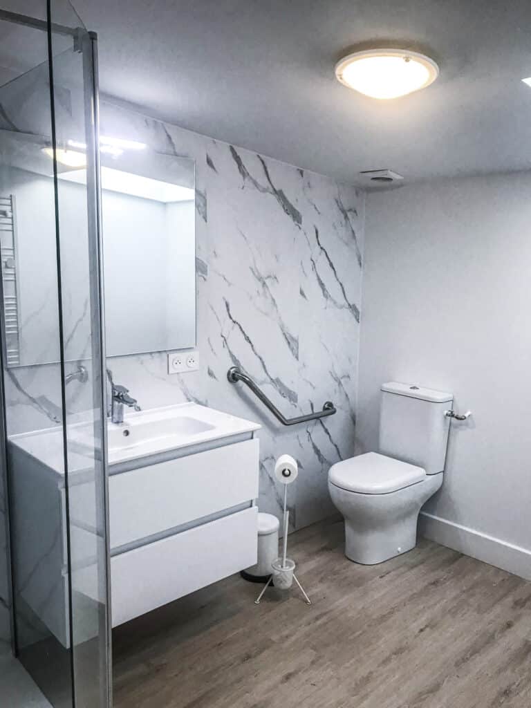 Salle de bain moderne lumineuse avec douche vitrée, meuble lavabo blanc, WC, barre d'appui murale effet marbre et sol bois clair.