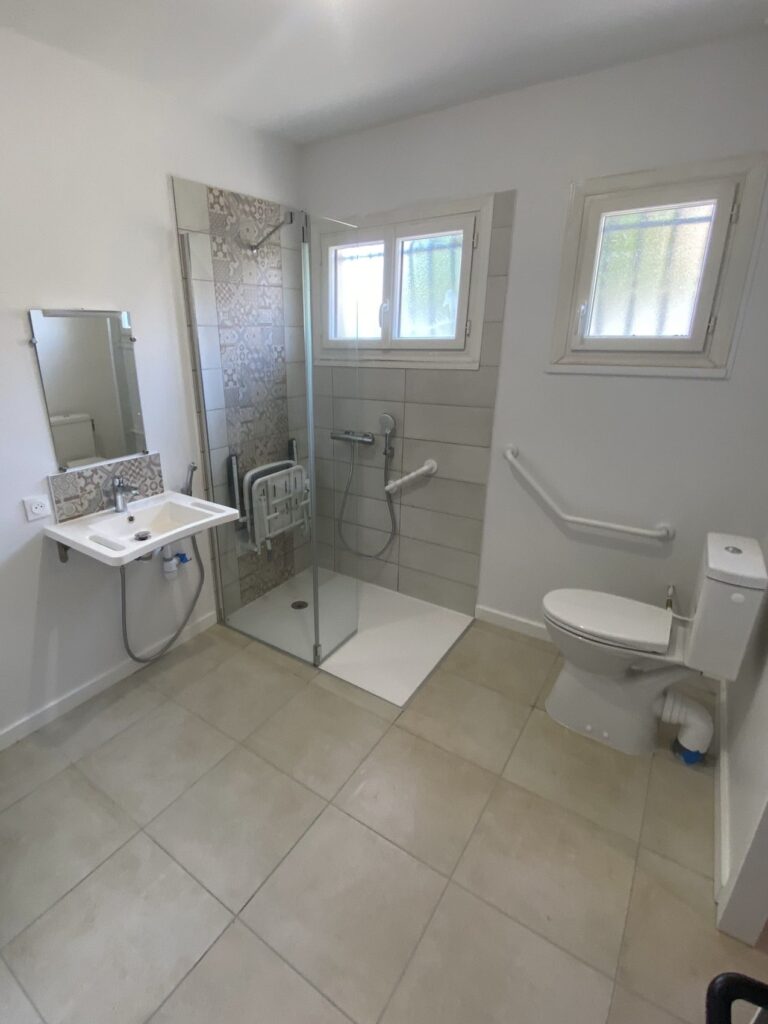 Salle de bain PMR moderne et lumineuse avec lavabo mural, miroir, douche à l'italienne, siège rabattable, barres d'appui et WC. Tons clairs.