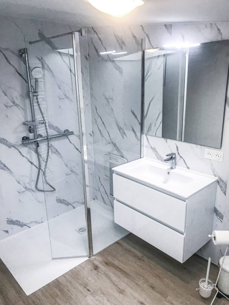 Salle de bain moderne : douche à l'italienne, murs effet marbre blanc, meuble vasque flottant blanc et miroir. Prises 250V.