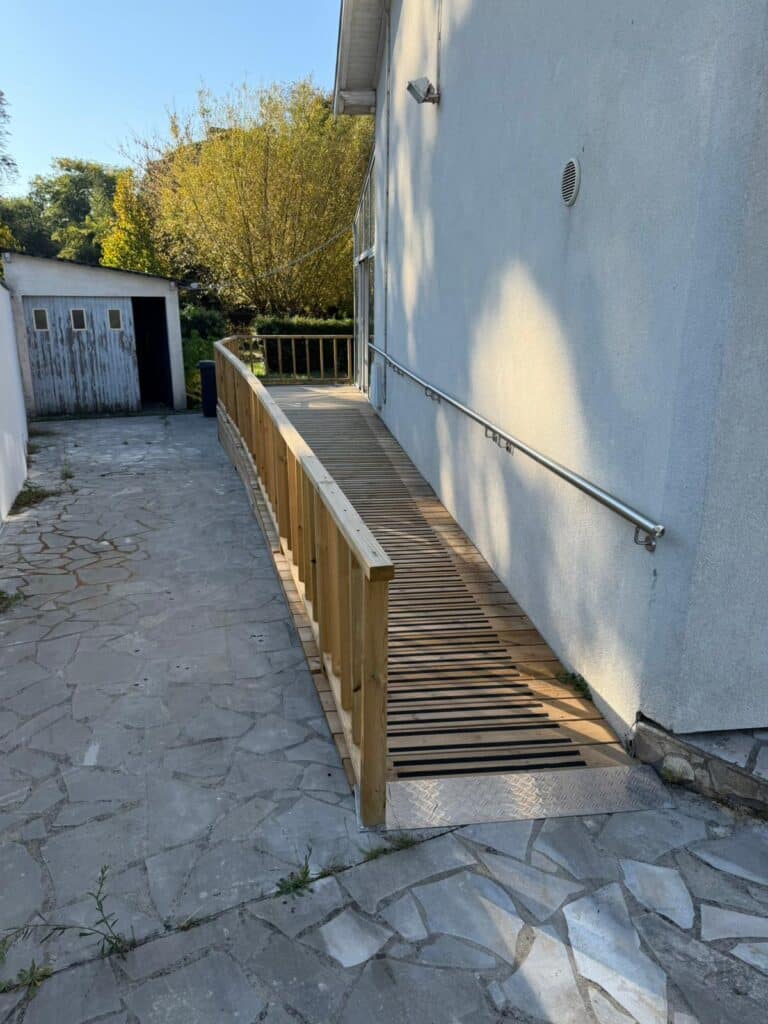 Rampe d'accès PMR en bois avec lattes antidérapantes noires, main courante inox murale et garde-corps en bois, installée près d'un chemin de pierres.