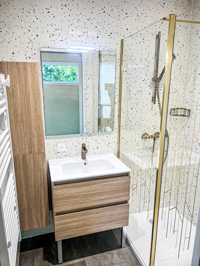 Salle de bain moderne avec meuble vasque bois, carrelage terrazzo et douche italienne aux finitions dorées.