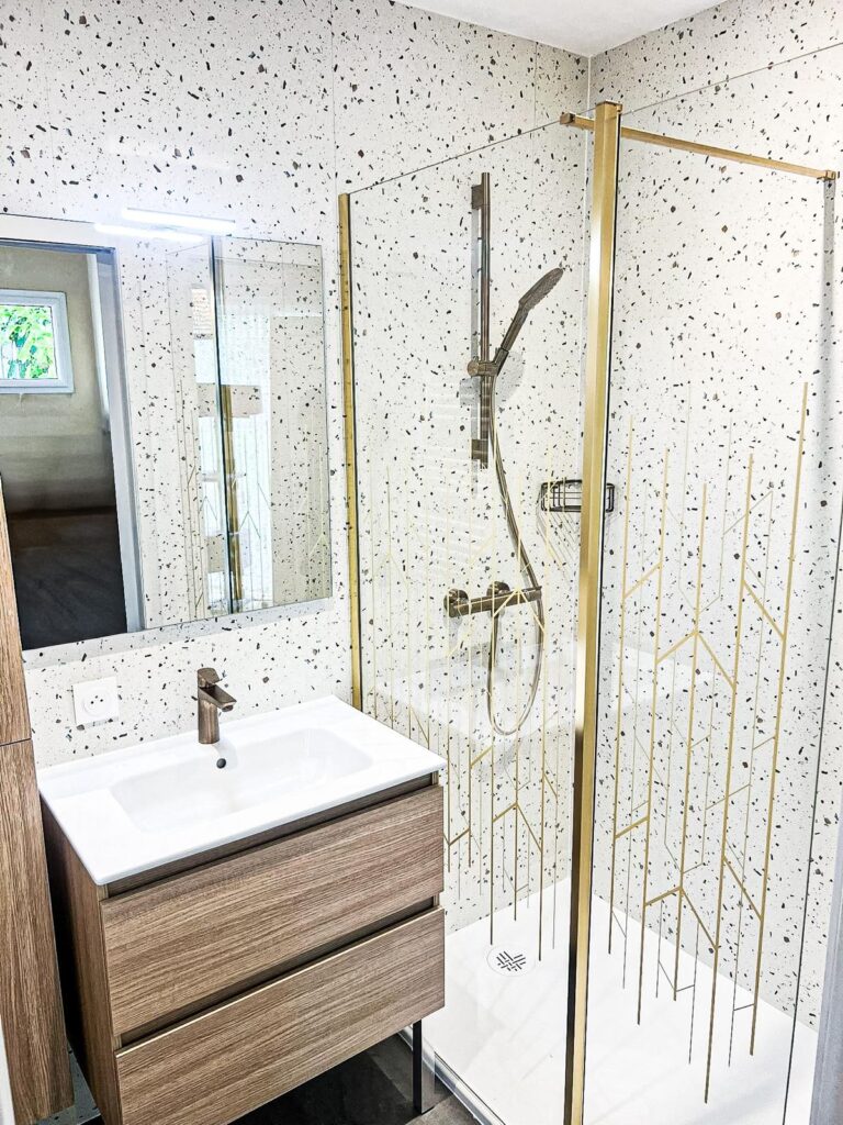 Salle de Bain Moderne : Style Terrazzo et Détails Dorés Salle de bain moderne avec murs terrazzo, meuble vasque en bois et douche vitrée à motifs géométriques dorés.